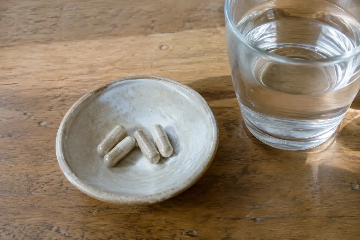 Quelques gélules beiges dans une coupelle en céramique, à côté d'un verre d'eau